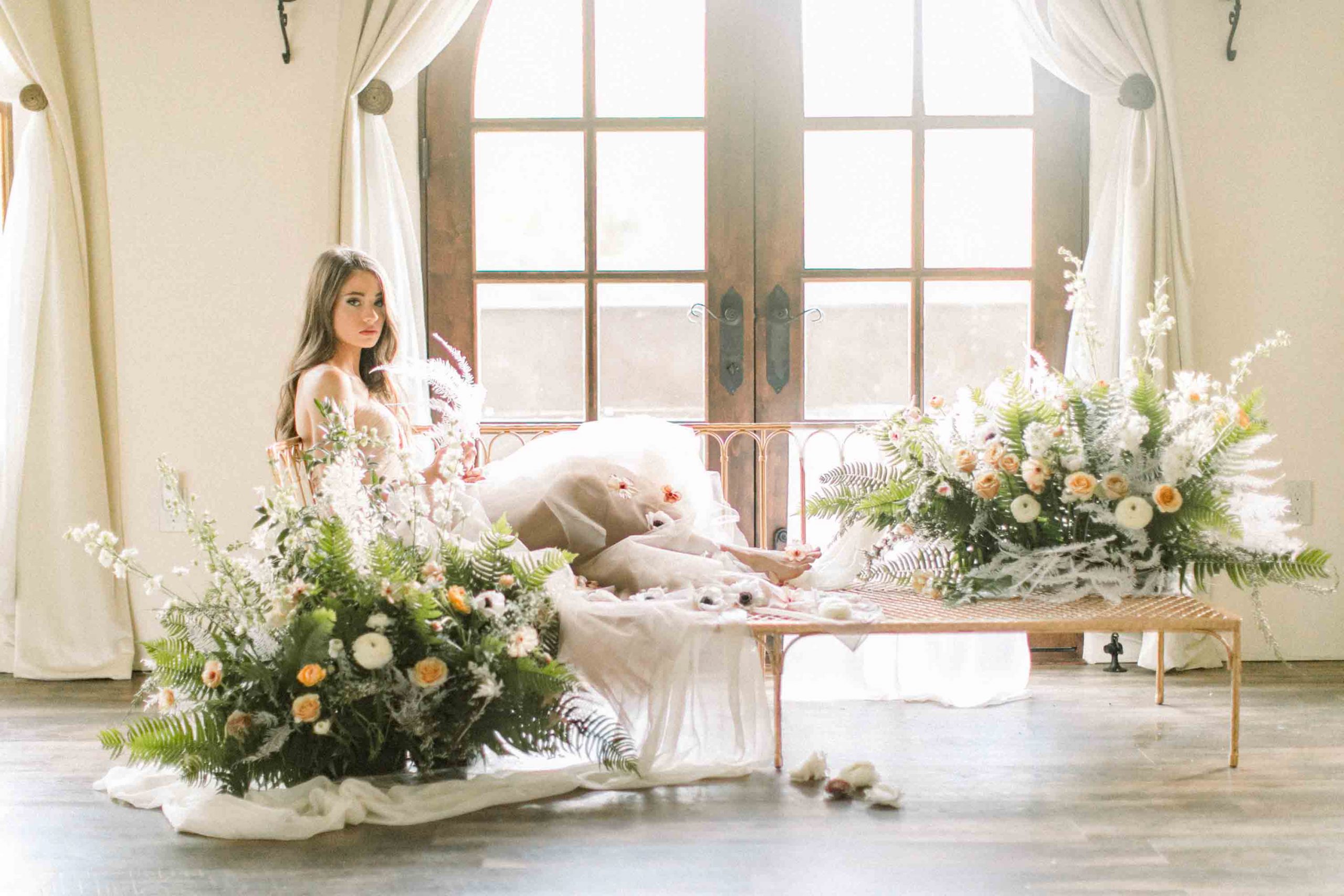 Elegant bridal portrait with soft white florals and greenery in a sunlit indoor setting.