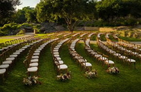 Curved rows of wooden wedding chairs with floral aisle arrangements in a garden setting.