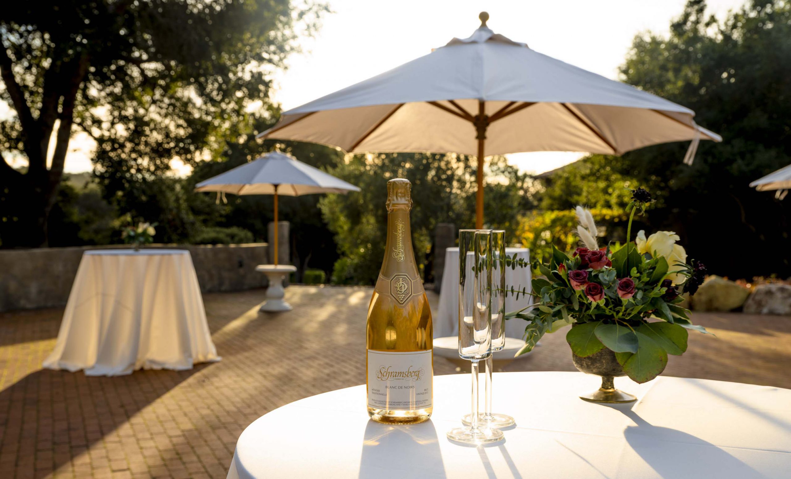 Outdoor wedding cocktail area with champagne bottle, floral arrangement, and white umbrellas at sunset.