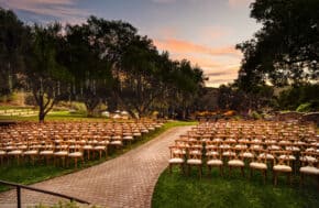 Outdoor wedding ceremony setup with rows of wooden chairs facing a garden aisle at sunset.