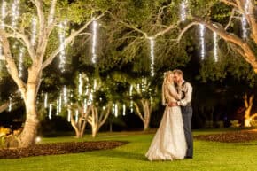 Bride and groom sharing a kiss under twinkling lights hanging from trees.